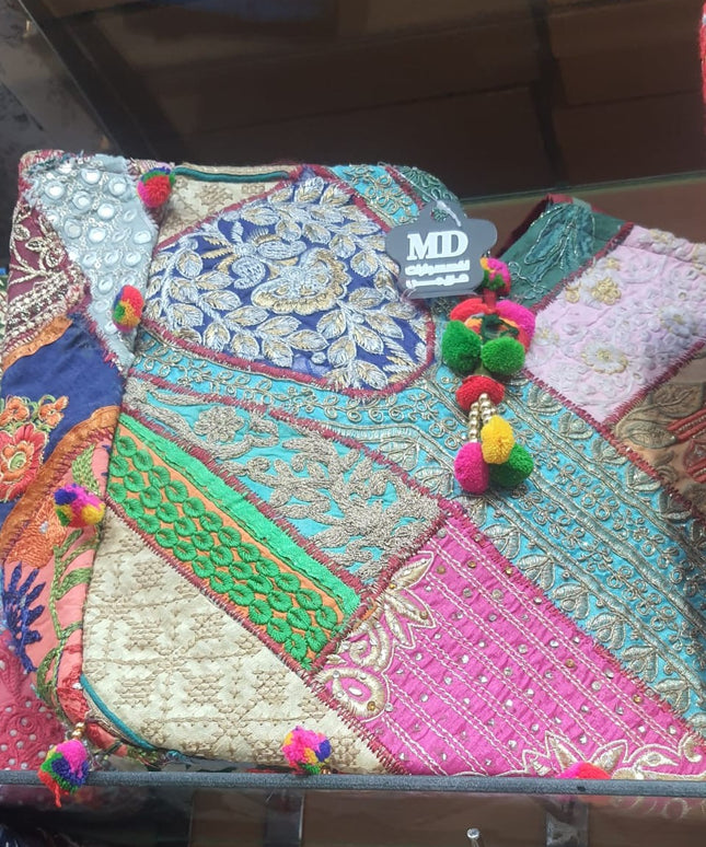 صورة مقربة لشنطة حديقة القصر تظهر دقة فن الباتشورك التراثي - دائرة مركزية كبيرة بزخارف بيزلي ملكية مطرزة بالخيوط الذهبية المعدنية على حرير أزرق ملكي محاطة بـ15-20 قطعة قماشية مختلفة بألوان متناسقة: أخضر، وردي، بيج، بنفسجي - كل قطعة مطرزة بنمط فريد بخيوط Zari الذهبية والفضية مع مرايا Shisha صغيرة لامعة وخرز ملون - 20-25 شراشيب بوم بوم صوفية ملونة تحيط بالشنطة - حزام كتف عريض مطرز ومبطن - تحفة فنية تراثية عملية من كنوز مصر
