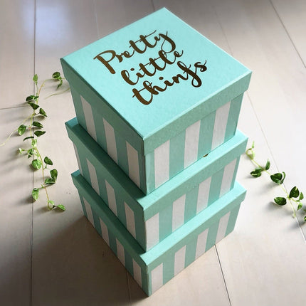 طقم صناديق Pretty Little Things التيفاني الأخضر النعناعي من كنوز مصر مكدس بثلاثة أحجام متدرجة على أرضية فاتحة محاطاً بنباتات خضراء طبيعية متدلية، يظهر الاستخدام المتعدد للصناديق كتغليف وتخزين وديكور مع كتابة Pretty Little Things الذهبية والخطوط البيضاء الكانديز بوضوح كامل

